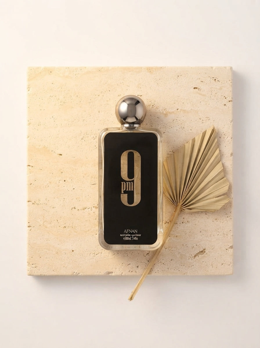 A charcoal black perfume bottle sits on a warm cream surface with soft golden lighting and a decorative dried leaf.